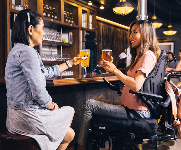 dos mujeres sonriendo y tomando una cerveza en un bar, una de ellas usa silla de ruedas